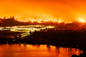 Forest fires encroaching on a town, reflected in the water.