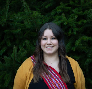 Headshot of Erin Blondeau, a light-skinned woman with dark hair wearing a dark yellow cardigan and a red ceinture fléchée (Métis sash), standing against a background of evergreens.